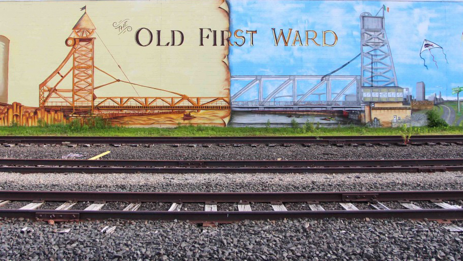 A mural reads "Old First Ward" beyond sets of rustic railways.